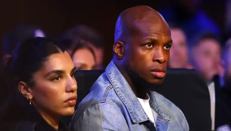 Man in a light denim jacket seated in a dimly lit audience, looking toward the stage; a woman with earrings sits beside him in the foreground.