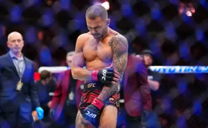 Shirtless UFC fighter with a tattooed left arm stands inside the cage, wearing red and black shorts and UFC gloves, looking down in focus.