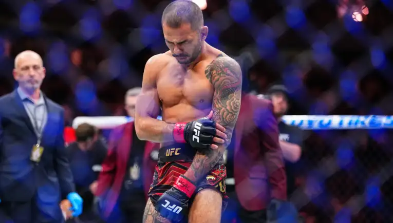 Shirtless UFC fighter with a tattooed left arm stands inside the cage, wearing red and black shorts and UFC gloves, looking down in focus.