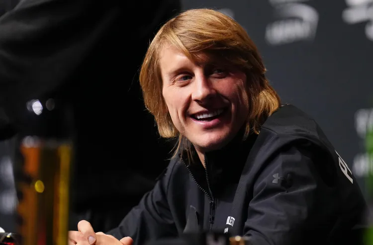 Smiling man with shoulder-length blond hair sits at a table during a panel, wearing a black jacket.