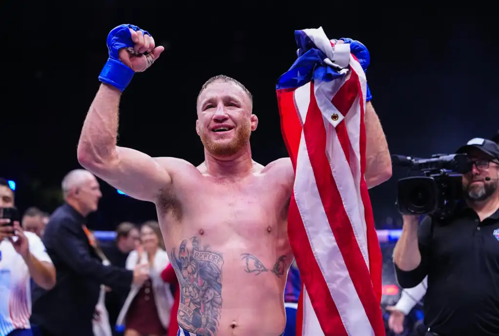 Boxer with blue gloves raises his fists in victory, wrapped in a British flag, amid a crowded arena and cameras.
