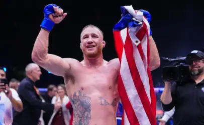 Boxer with blue gloves raises his fists in victory, wrapped in a British flag, amid a crowded arena and cameras.