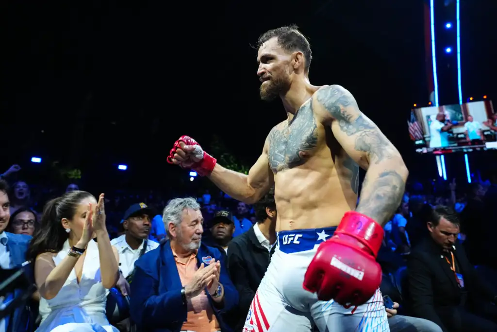 Male UFC fighter shirtless with tattoos and red gloves entering a crowded arena at night, fans cheering around him.