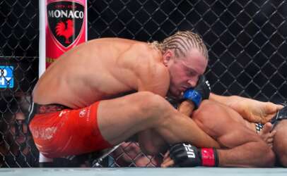 Two UFC fighters grapple on the mat inside a chain-link cage; one in red shorts pins the other with blue gloves visible.
