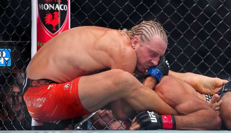 Two UFC fighters grapple on the mat inside a chain-link cage; one in red shorts pins the other with blue gloves visible.