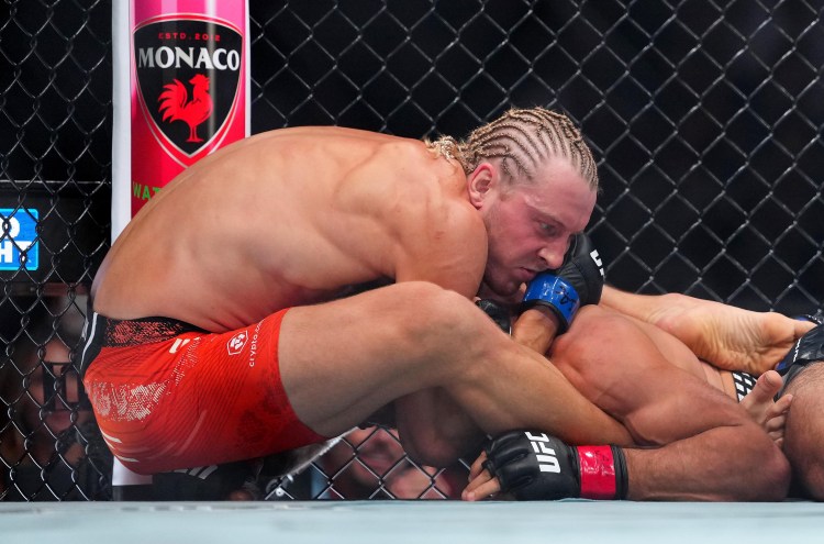 Two UFC fighters grapple on the mat inside a chain-link cage; one in red shorts pins the other with blue gloves visible.