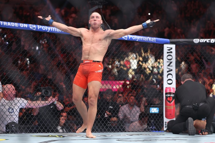 MMA fighter in orange shorts stands in the octagon with arms raised, crowd cheering behind him and a Monaco-branded post nearby, celebrating a victory.