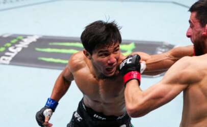 Two shirtless UFC fighters exchange a punch in the octagon, with one fighter landing a punch to the other's face while both wear UFC gloves.
