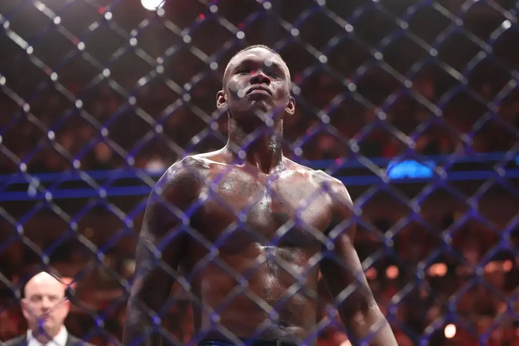 Male mixed martial artist stands shirtless inside a chain-link cage, eyes forward, sweat glistening under arena lights.