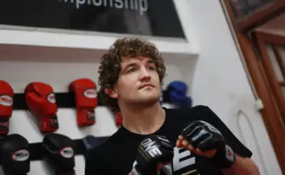 Young man with curly hair wearing boxing gloves in a gym, standing with fists up near a wall of red gloves and a banner reading Championship (partial).