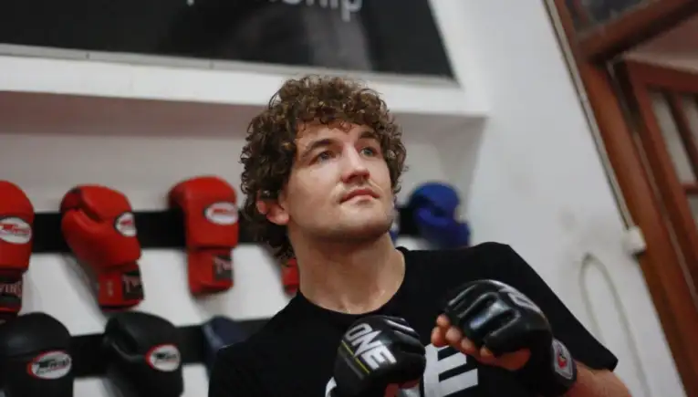Young man with curly hair wearing boxing gloves in a gym, standing with fists up near a wall of red gloves and a banner reading Championship (partial).