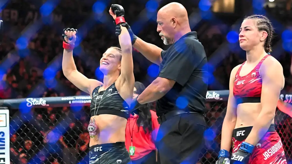 Female UFC fighter in camo shorts raises both arms in victory as the referee declares her winner inside the cage; opponent in red sports bra stands nearby with a somber expression.
