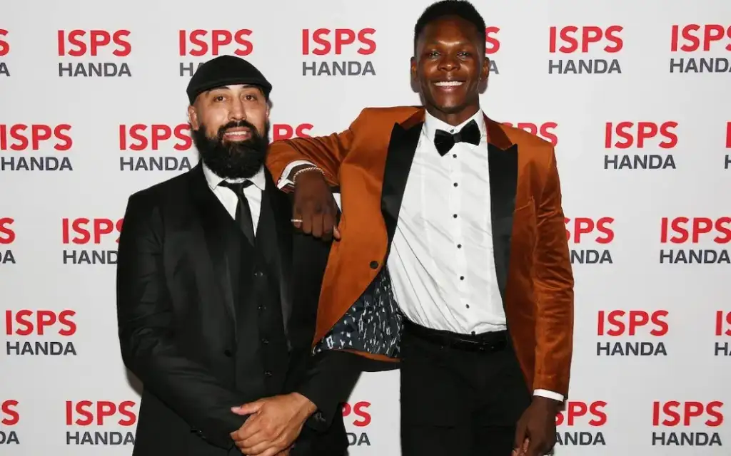 Two men in formal attire pose on a red-carpet backdrop with ISPS HANDA logos; left man in black suit and cap, right man in an orange blazer and bow tie.