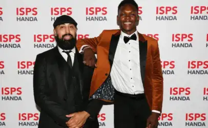 Two men in formal attire pose on a red-carpet backdrop with ISPS HANDA logos; left man in black suit and cap, right man in an orange blazer and bow tie.