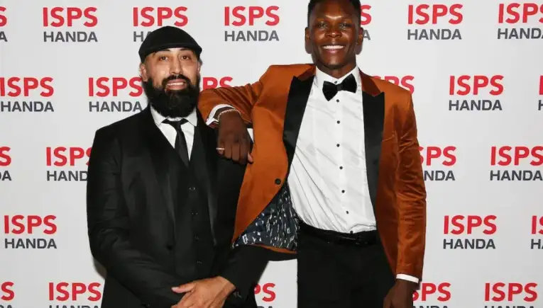 Two men in formal attire pose on a red-carpet backdrop with ISPS HANDA logos; left man in black suit and cap, right man in an orange blazer and bow tie.