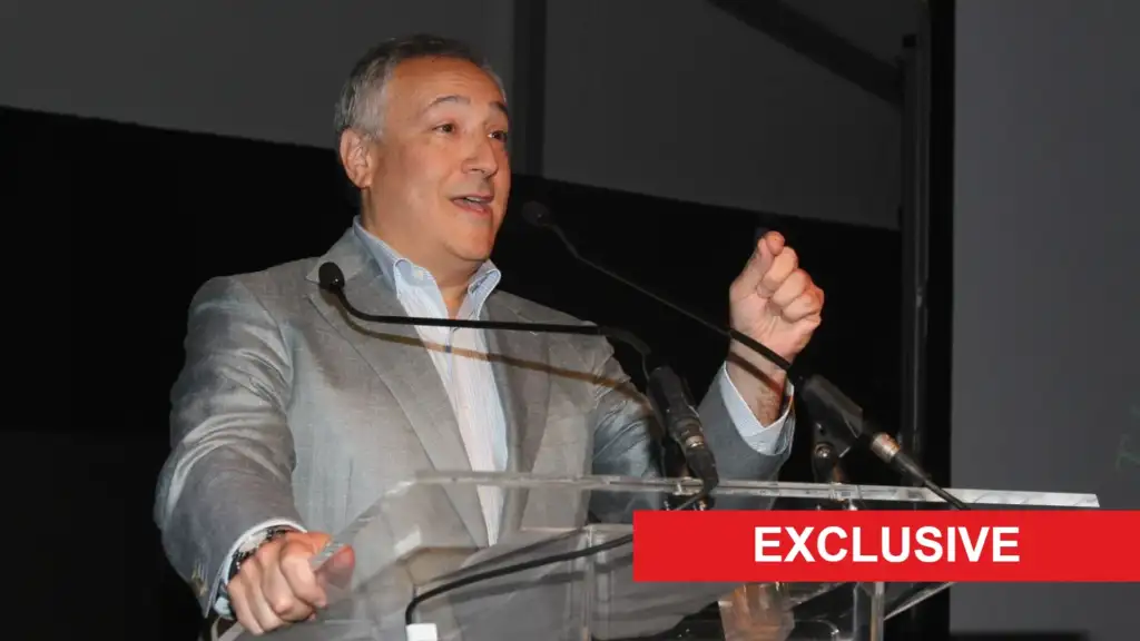 Man in a gray suit giving a speech at a clear podium on stage, with a red EXCLUSIVE banner across the bottom-right.