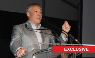 Man in a gray suit giving a speech at a clear podium on stage, with a red EXCLUSIVE banner across the bottom-right.