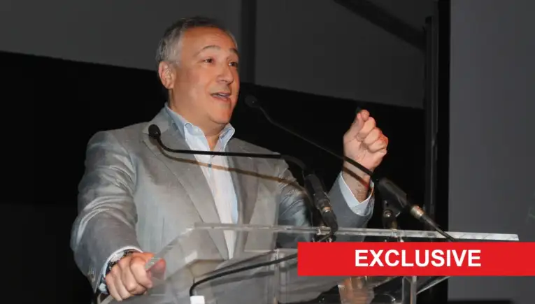 Man in a gray suit giving a speech at a clear podium on stage, with a red EXCLUSIVE banner across the bottom-right.