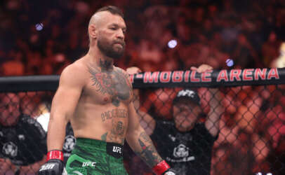 MMA fighter with tattoos and a trimmed beard stands in the UFC octagon, wearing green shorts and red gloves, with the crowd and a banner reading 'MOBILE ARENA' in the background.