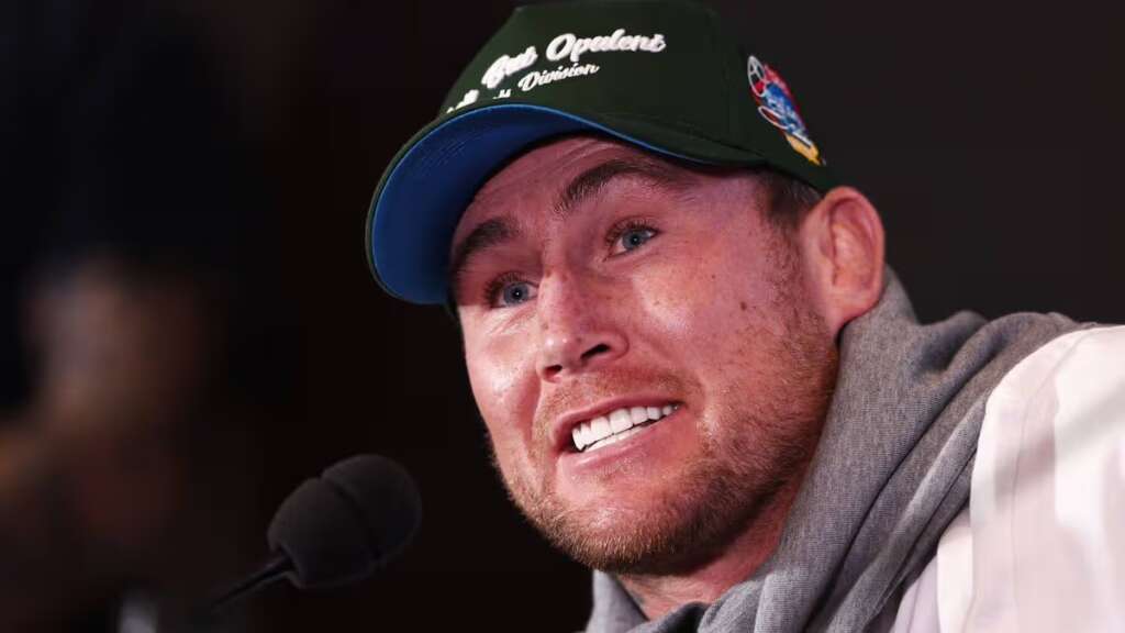 Close-up of a man wearing a green cap with logos, speaking into a microphone with a friendly expression. The brim is blue, and he has a light beard and a gray scarf over a white jacket.