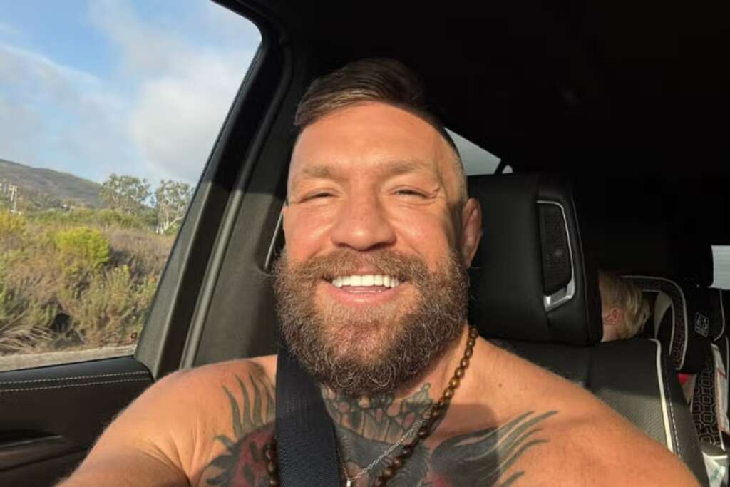 Selfie of a smiling man with a beard and chest tattoos in the front seat of a car, wearing a seatbelt as sunlight falls on his face.