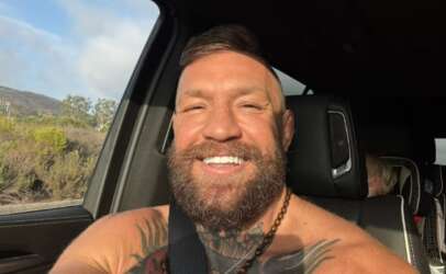 Selfie of a smiling man with a beard and chest tattoos in the front seat of a car, wearing a seatbelt as sunlight falls on his face.