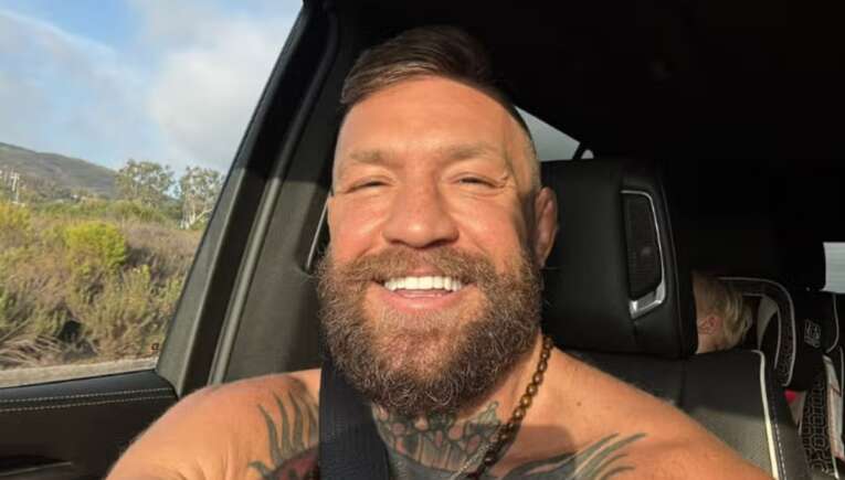 Selfie of a smiling man with a beard and chest tattoos in the front seat of a car, wearing a seatbelt as sunlight falls on his face.