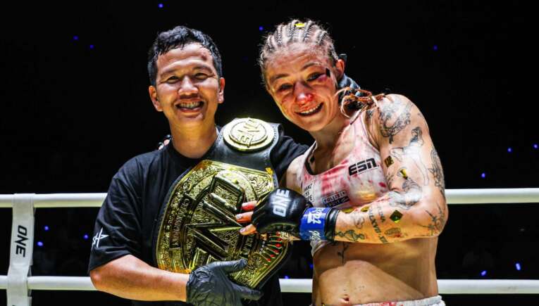 Two fighters pose in a ring, one wearing a gold championship belt and smiling with his arm around a teammate.