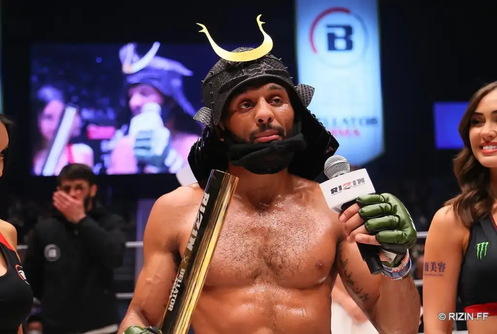 Boxer in a horned helmet being interviewed, holding a gold trophy and microphone in a ring. Green gloves, sweaty torso, crowd in the background.