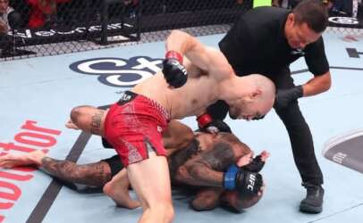 Two mixed martial arts fighters grapple on the mat as a referee watches from the side.