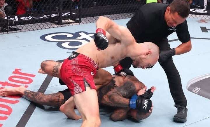 Two mixed martial arts fighters grapple on the mat as a referee watches from the side.
