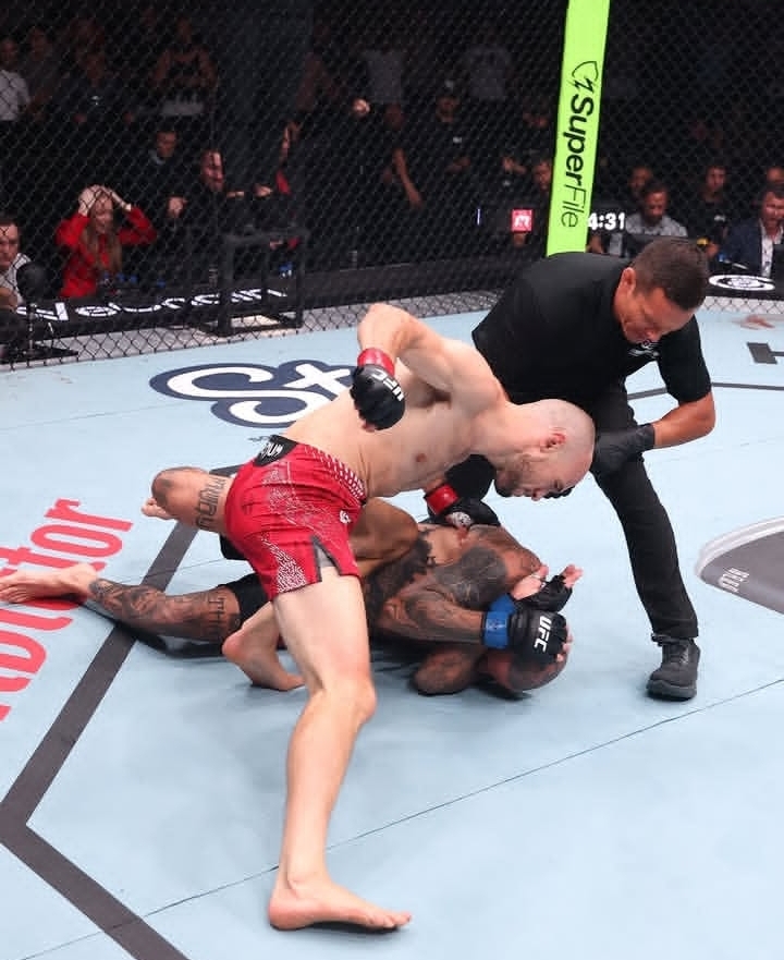 Two mixed martial arts fighters grapple on the mat as a referee watches from the side.