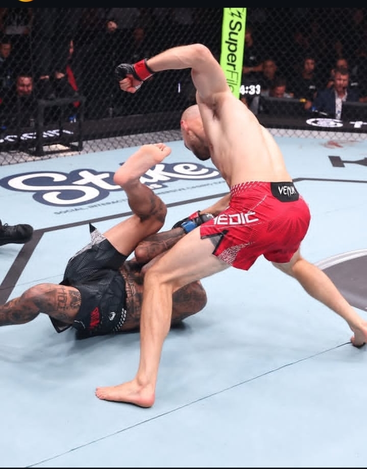 MMA fighters in a cage; one in red shorts delivering a knee/strike while the other lies on the canvas in black shorts amid a grappling exchange.