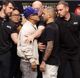 Two male fighters in a tense face-off at a UFC event, surrounded by staff and security nearby.