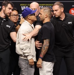 Two male fighters in a tense face-off at a UFC event, surrounded by staff and security nearby.