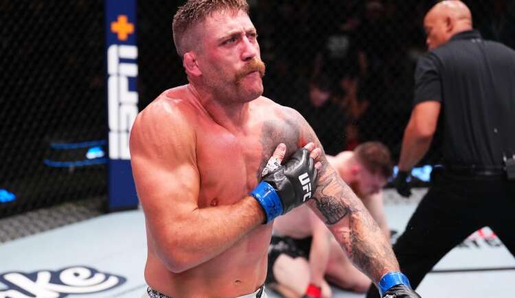 Muscular UFC fighter with blue gloves and tattoos crosses his arms over his chest inside the octagon during a bout.