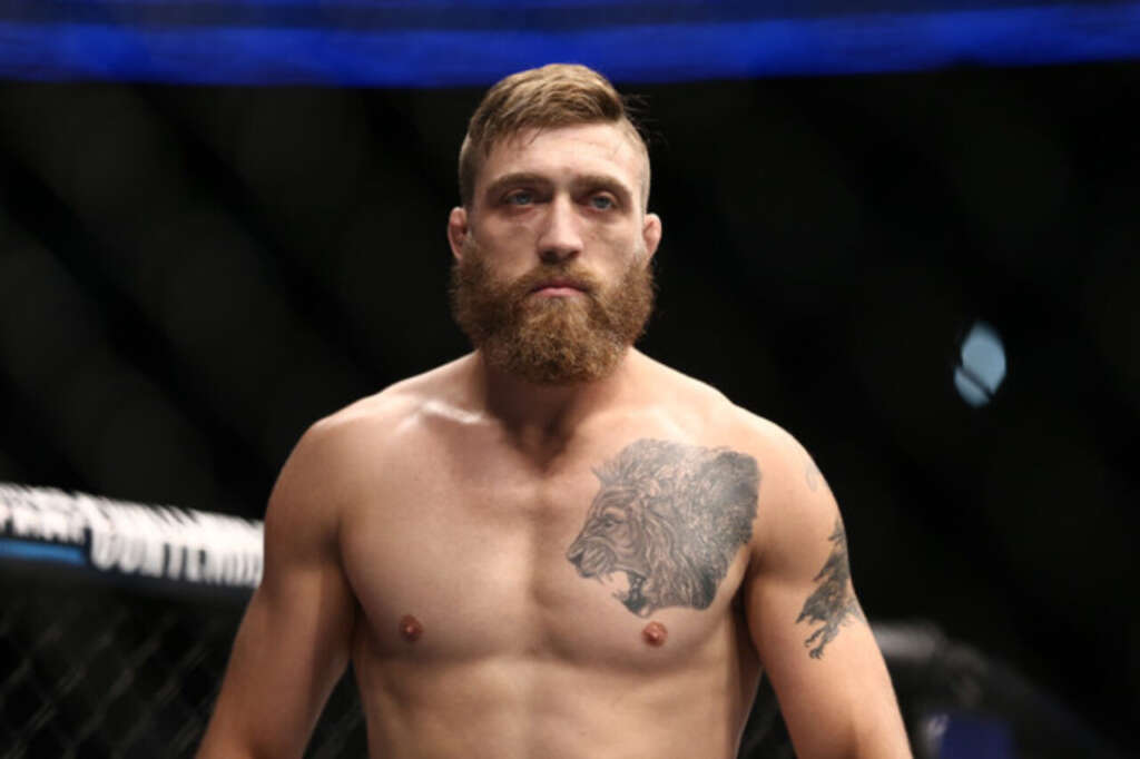 Bearded male MMA fighter, shirtless, standing inside a fenced octagon, with a large lion tattoo on his chest.