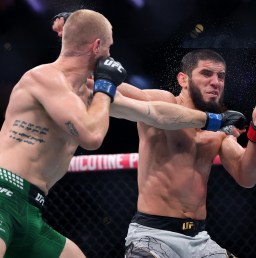 Two male mixed martial artists trade punches in a cage; fighter in green shorts throws a right toward the opponent in gray shorts wearing blue gloves.