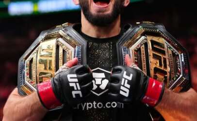 UFC fighter with two championship belts shouting in victory inside a crowded arena; gloves say UFC and crypto.com logo visible.