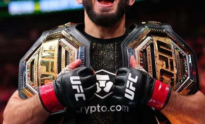 UFC fighter with two championship belts shouting in victory inside a crowded arena; gloves say UFC and crypto.com logo visible.