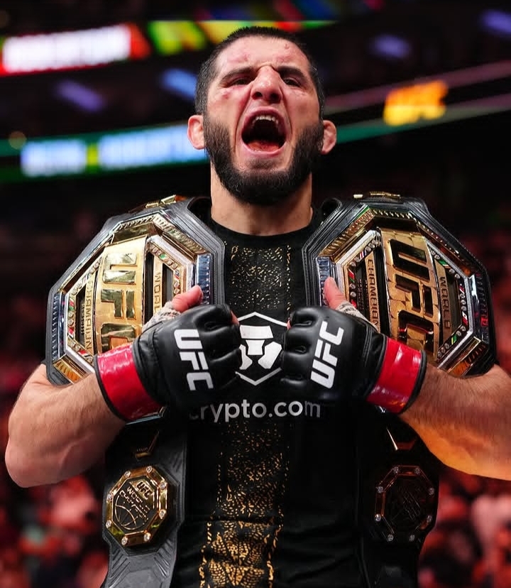 UFC fighter with two championship belts shouting in victory inside a crowded arena; gloves say UFC and crypto.com logo visible.