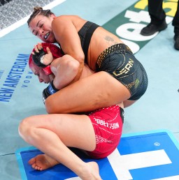 Two female mixed martial artists grapple on a blue competition mat, one pinning the other to the mat during a match still in progress.