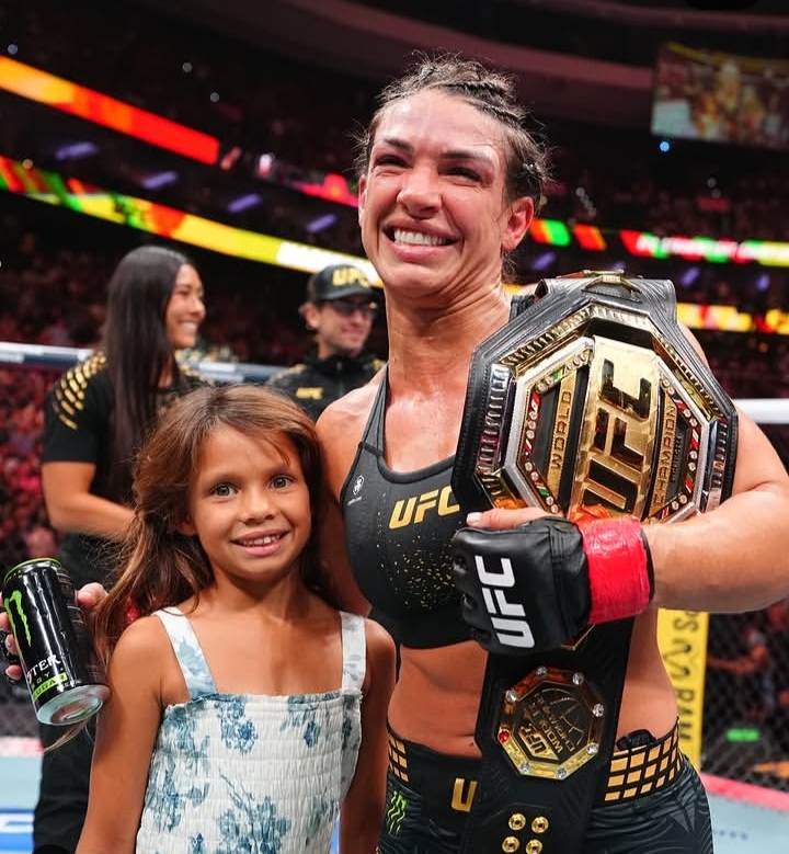 UFC fighter smiles with a championship belt over her shoulder, posing with a young girl in the arena.