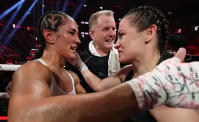 Two female boxers share a hug in the ring after a bout as their trainer smiles between them.