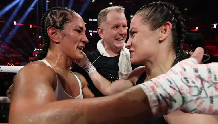 Two female boxers share a hug in the ring after a bout as their trainer smiles between them.