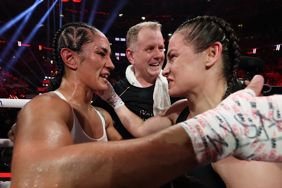 1001088802 - AfroCombat Two female boxers share a hug in the ring after a bout as their trainer smiles between them.
