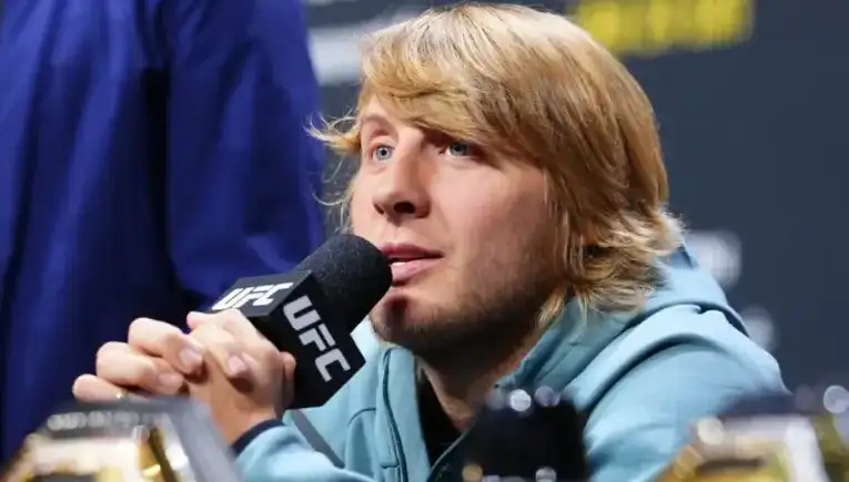 Blond-haired man speaking into a UFC microphone at a press conference with a focused expression.