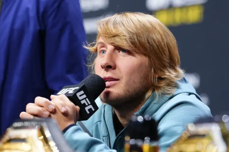 Blond-haired man speaking into a UFC microphone at a press conference with a focused expression.