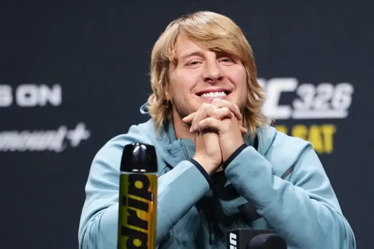 Young man with blond hair in a light blue hoodie smiles with hands clasped at a press conference, microphone in front of him and a yellow/black bottle nearby, against a dark backdrop.