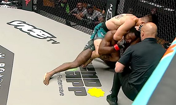 Two MMA fighters grapple on the canvas inside a cage; one in green trunks on top, the other underneath, with a referee nearby.